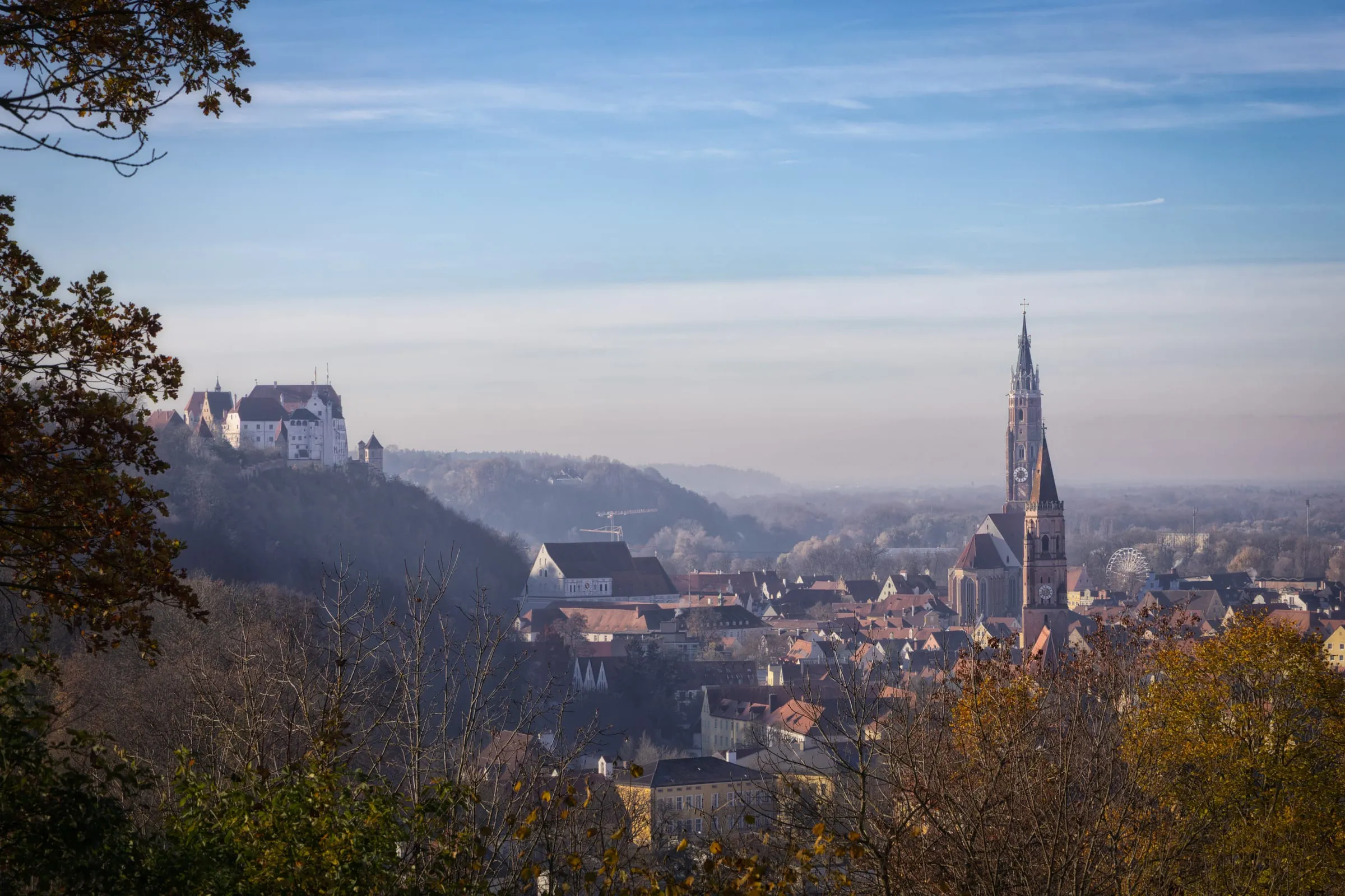 View through autumn foliage over Landshut, with Trausnitz Castle on a wooded hill to the left and St. Martin’s tower rising above the hazy old town.
