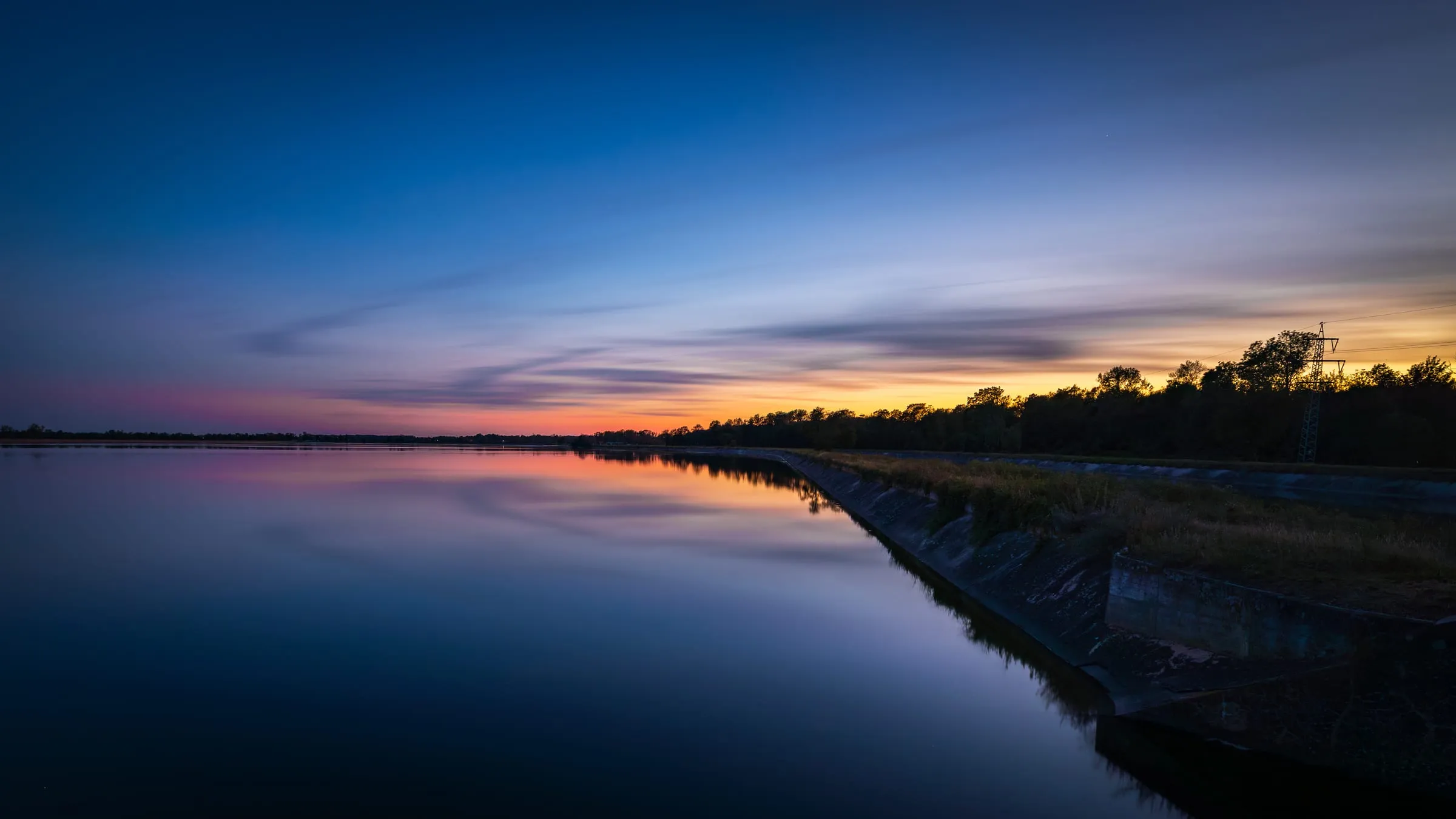 Nach Sonnenuntergang spiegelt der glatte Weiher den orangefarbenen Horizont und den tiefblauen Himmel; rechts verläuft ein Betondamm mit Bäumen und einem Strommast.