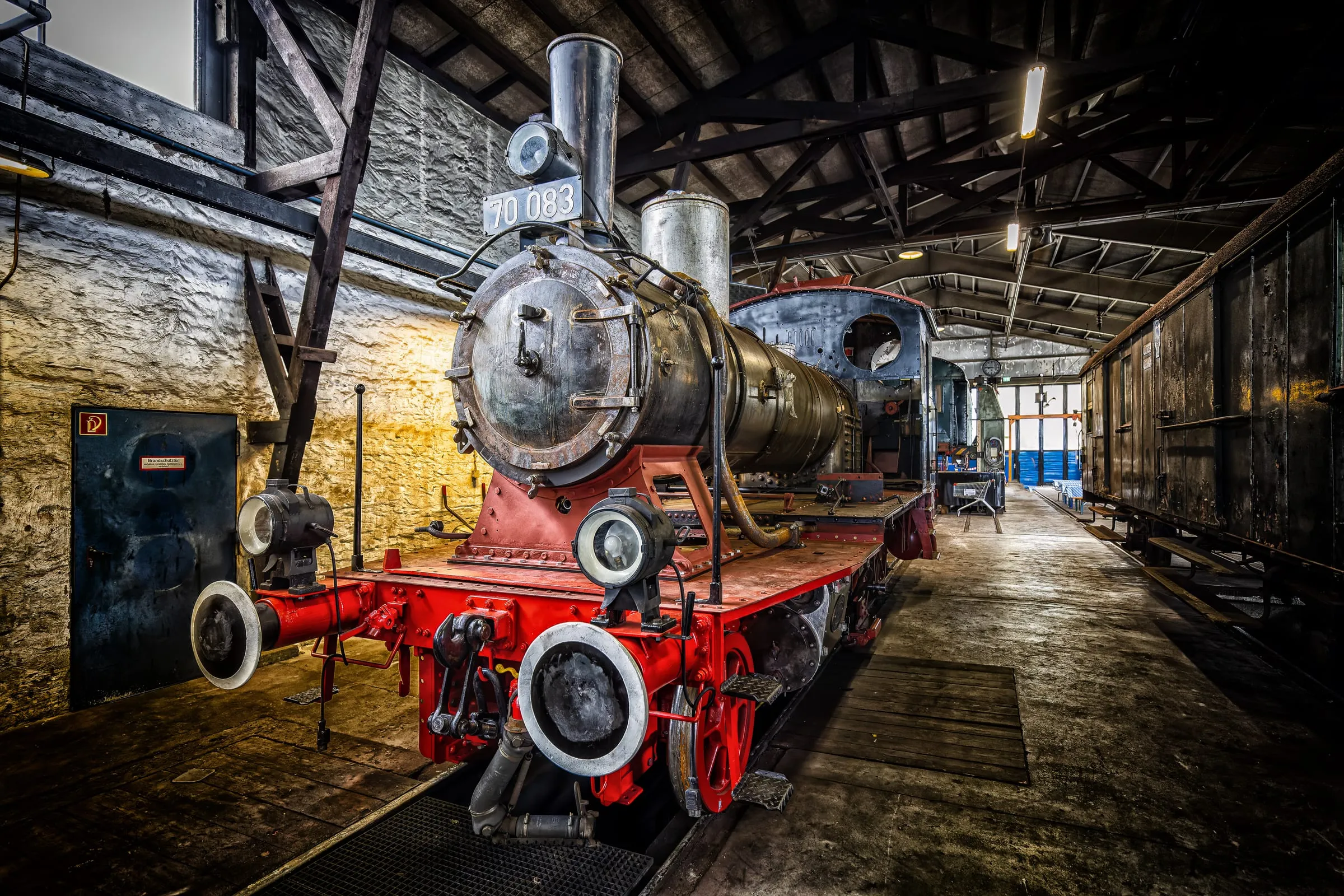 Steam locomotive 70 083 with a red frame and dark boiler in an engine shed with dark roof trusses and a stone wall, an old wagon on the right.
