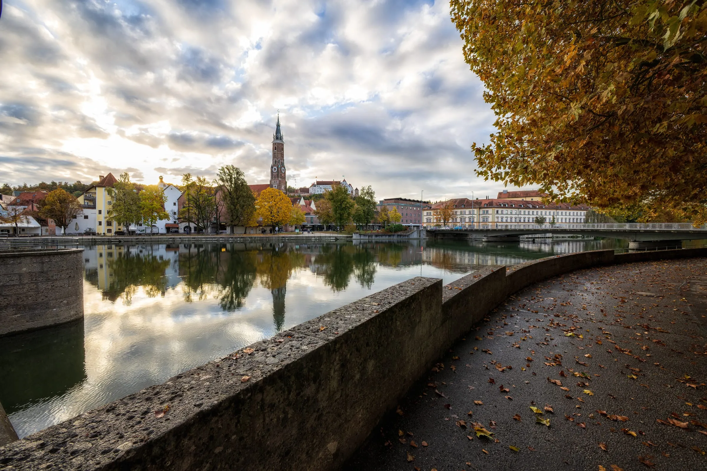 A curved river wall and leaf-strewn path along the Isar, St. Martin’s tower and trees reflected in the water, and a bridge on the right beneath a yellow autumn tree.