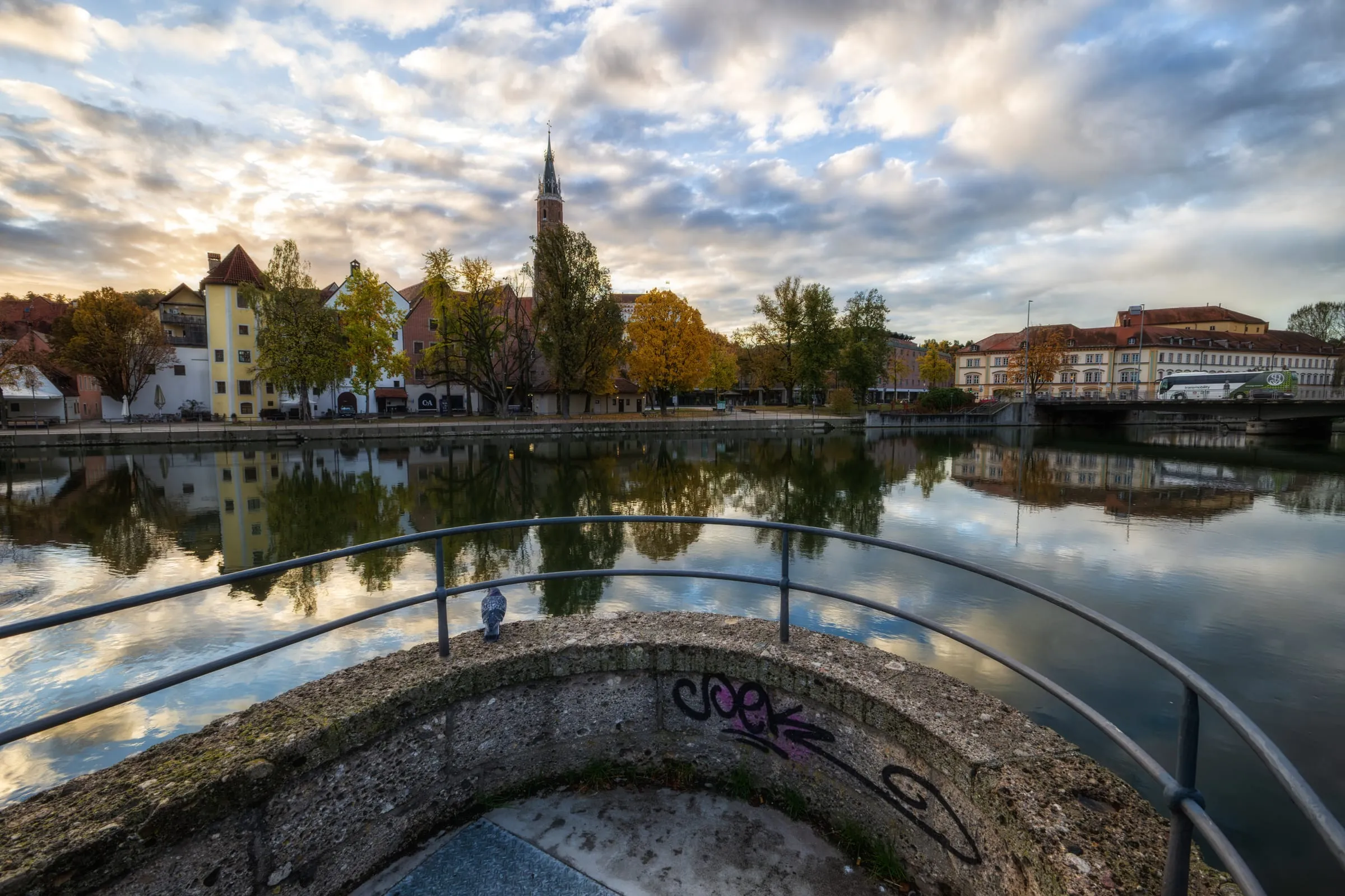 A round riverside platform with a railing and a pigeon; across the Isar, houses and autumn trees are reflected in the water, with St. Martin’s tower behind under clouds.