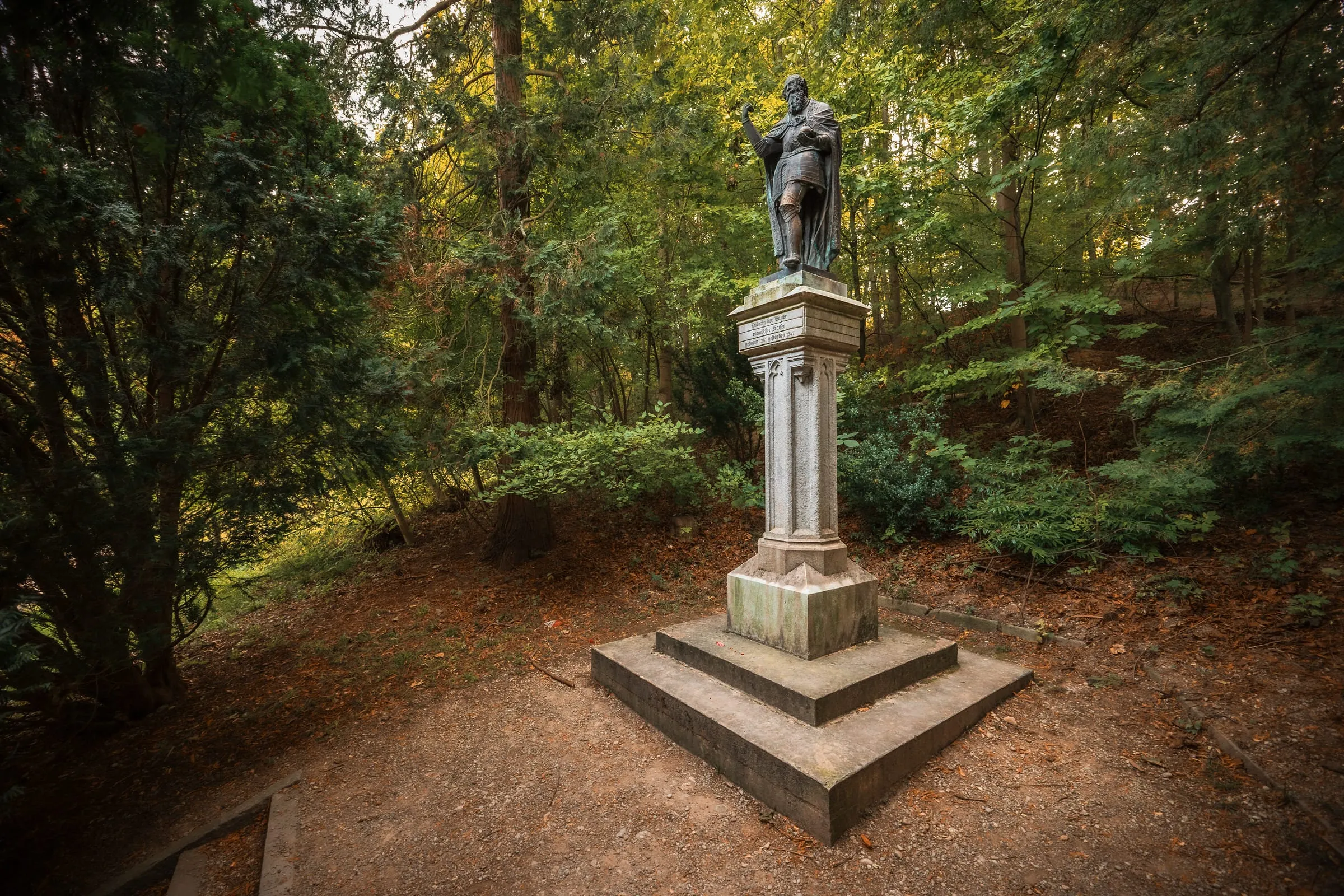Bronze statue of Ludwig the Bavarian on a tall inscribed stone pedestal, set on a leaf-covered clearing among green trees in the Hofgarten.