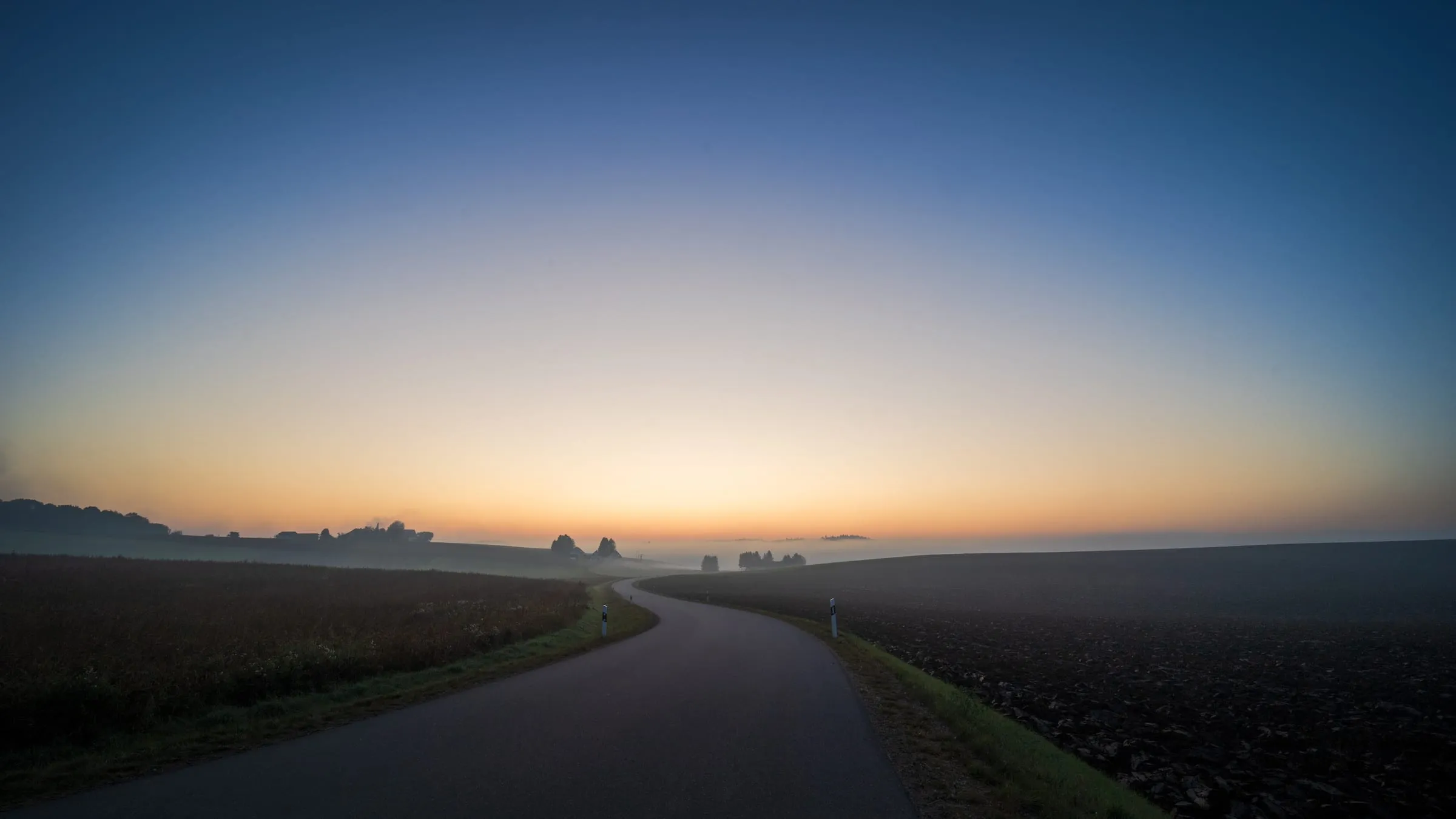 A narrow road curves between a field and ploughed farmland towards the horizon, which glows orange before sunrise, with fog lying in the hollows.