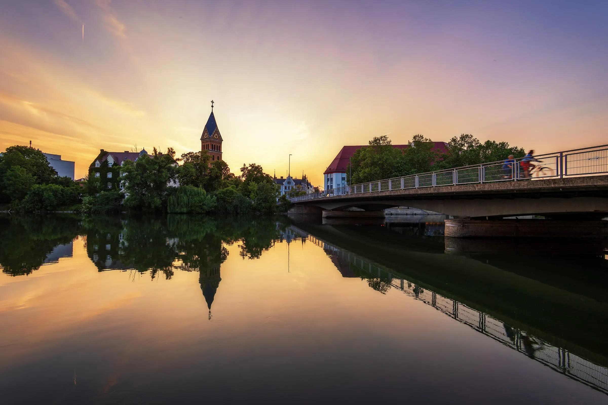 The still Isar reflects trees, a brick church tower and a bridge with two cyclists crossing, under an orange and violet sky at sunset.