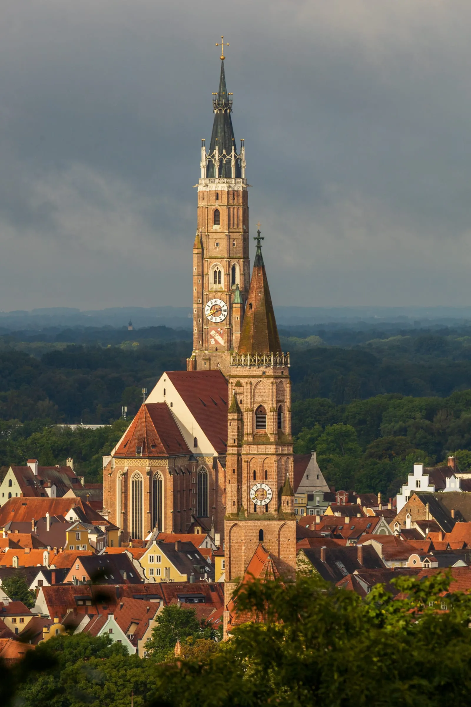 St. Martin’s brick tower lit by warm morning light against dark clouds, with a second tower and red old-town roofs in front and forest behind.
