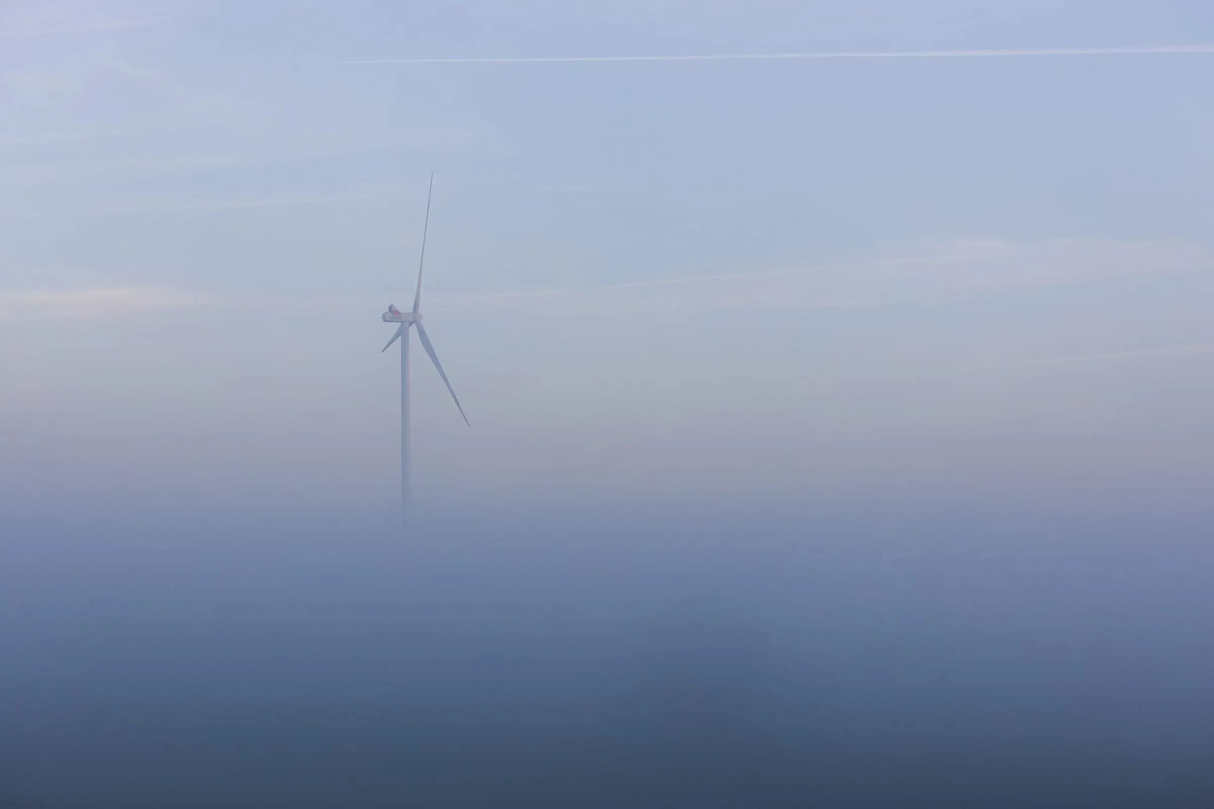 Ein einzelnes Windrad ragt links im Bild aus einer dichten Nebeldecke in den blassblauen Morgenhimmel, der von feinen Kondensstreifen durchzogen ist.