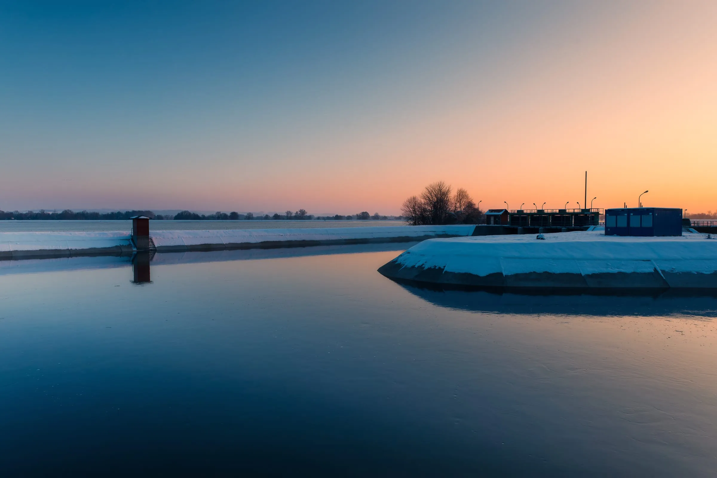 Dunkelblaue, glatte Wasserfläche zwischen verschneiten Betonufern, links ein rotes Häuschen im Wasser, rechts ein Bauwerk mit Laternen vor orangefarbenem Abendhimmel.